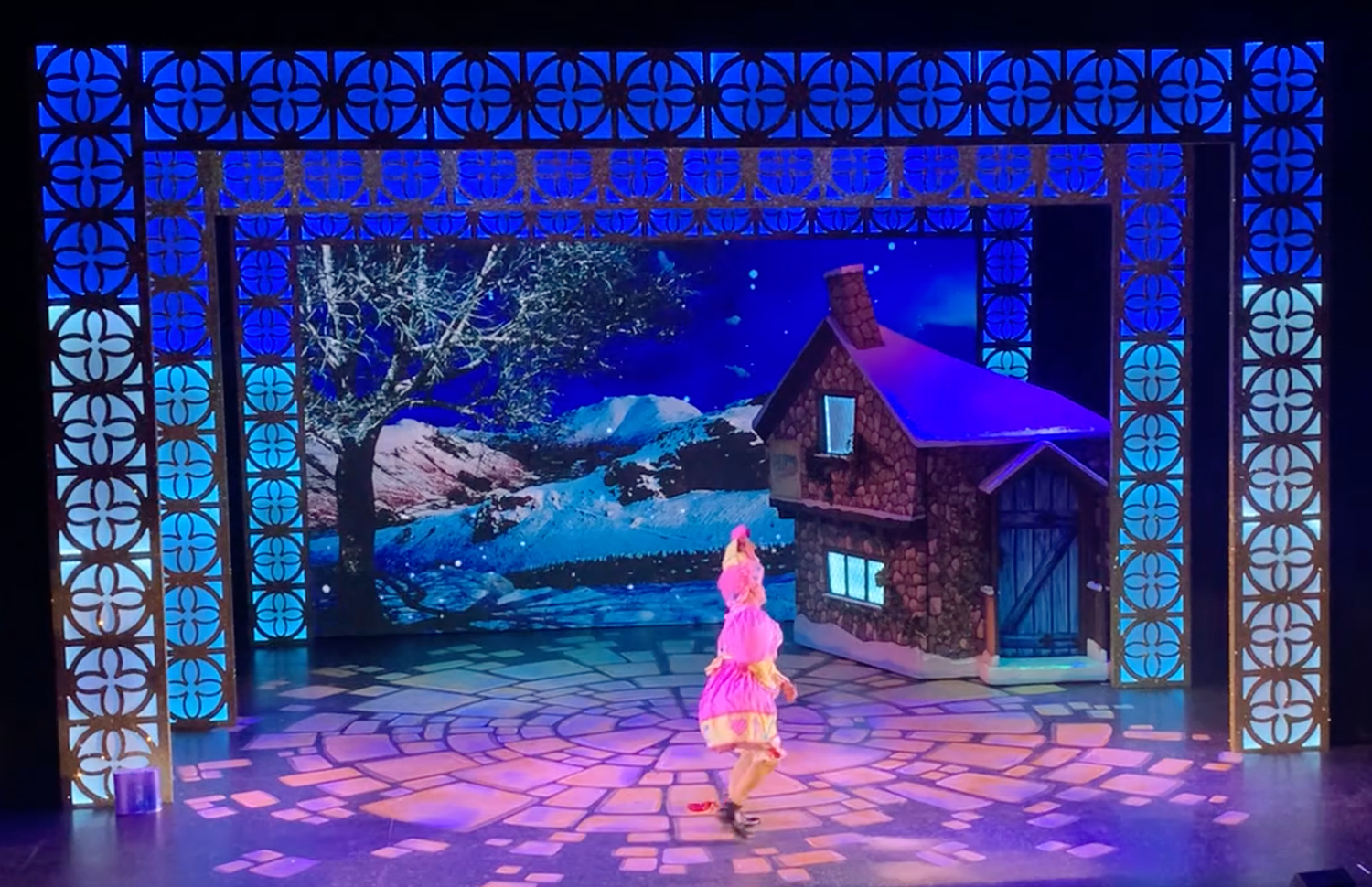Enchanting stage set with glowing blue geometric patterns and winter scenery featuring a dreamlike cottage backdrop.