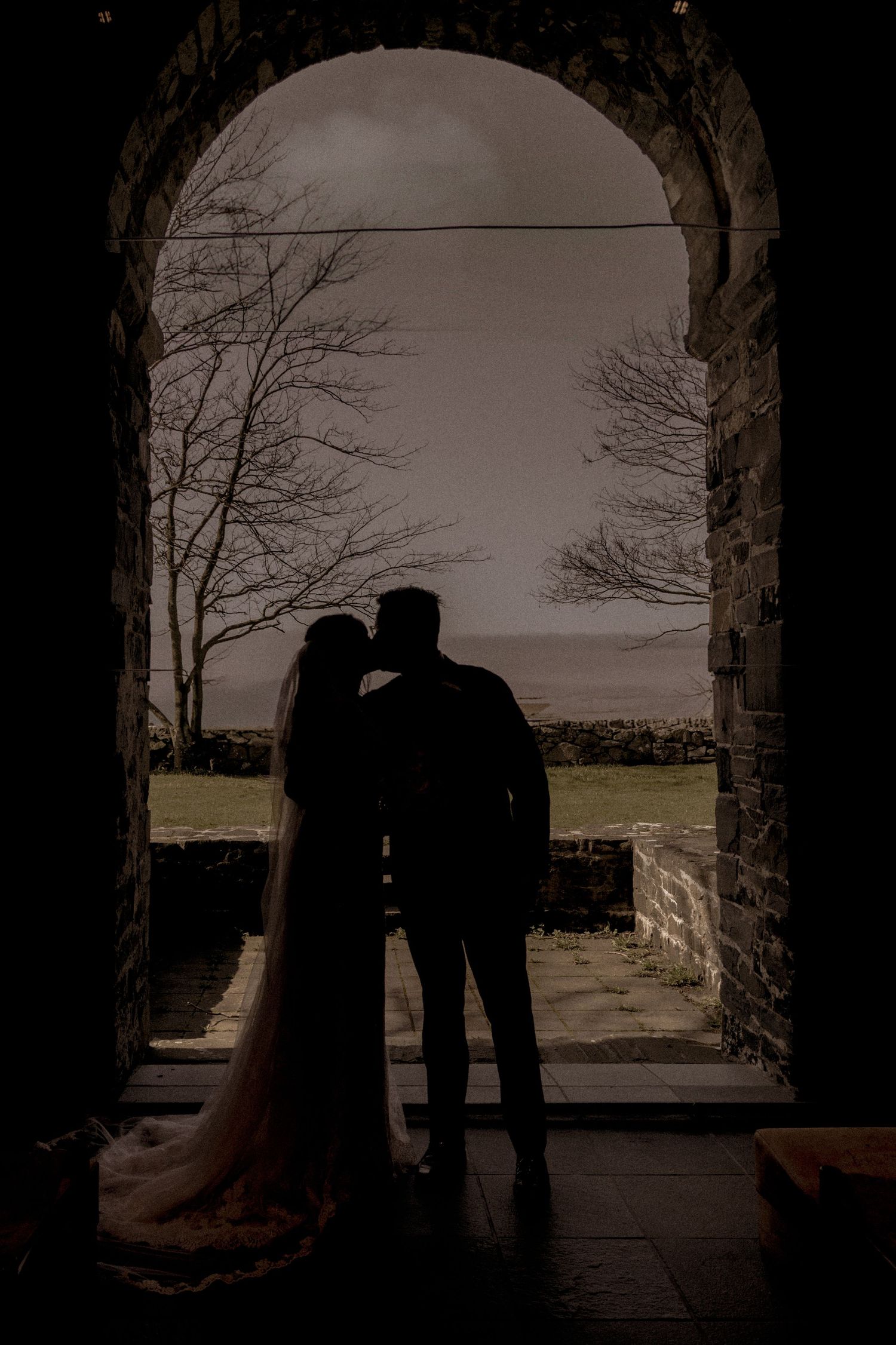 Silhouette of bride and groom kissing in stone archway with scenic outdoor backdrop.