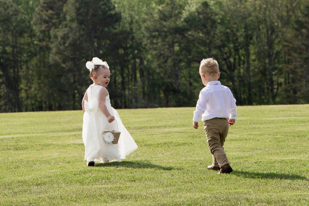 Young flower girl and ring bearer in formal wear run together going up the wedding aisle on a sunny green lawn and trees in the background.