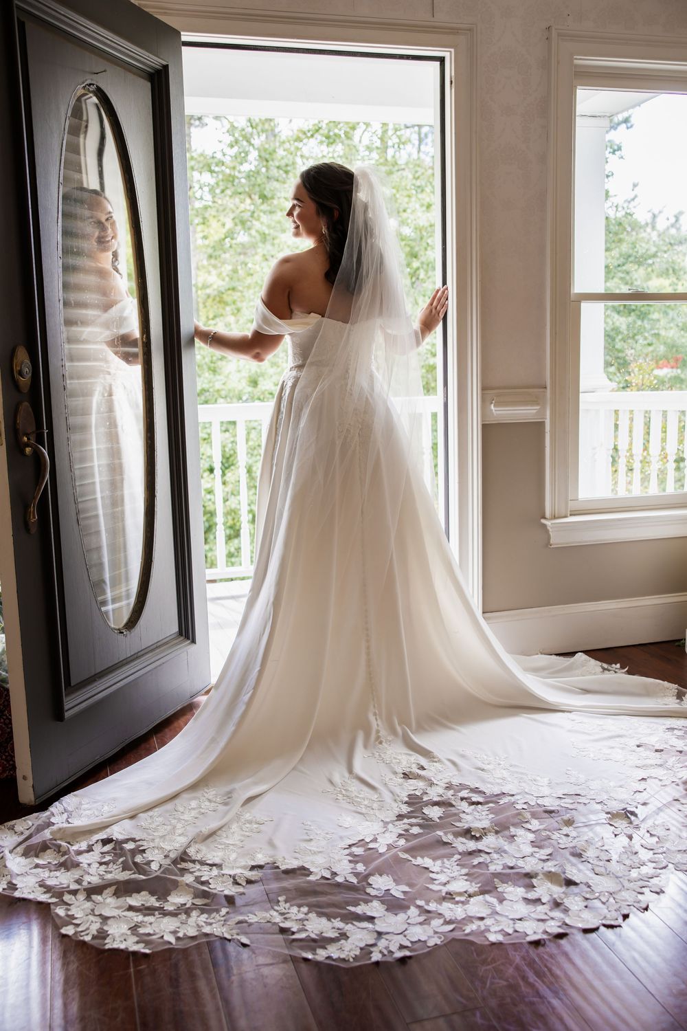 Bride in white strapless gown with long lace train looks at herself in oval mirror before wedding ceremony.