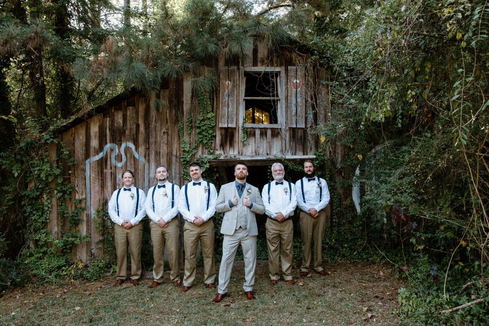 Groomsmen in light blue shirts and gray pants lean casually against a rustic wooden wall in an outdoor setting.