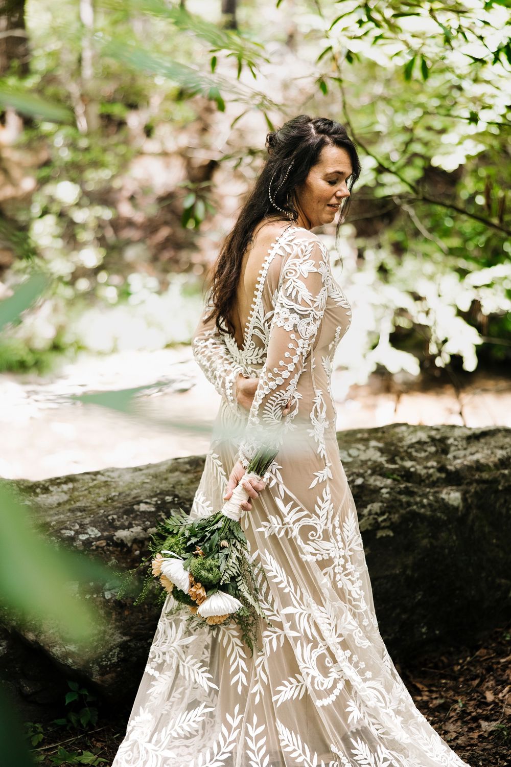Bride in lace boho wedding gown holding floral bouquet, posing in lush forest setting.