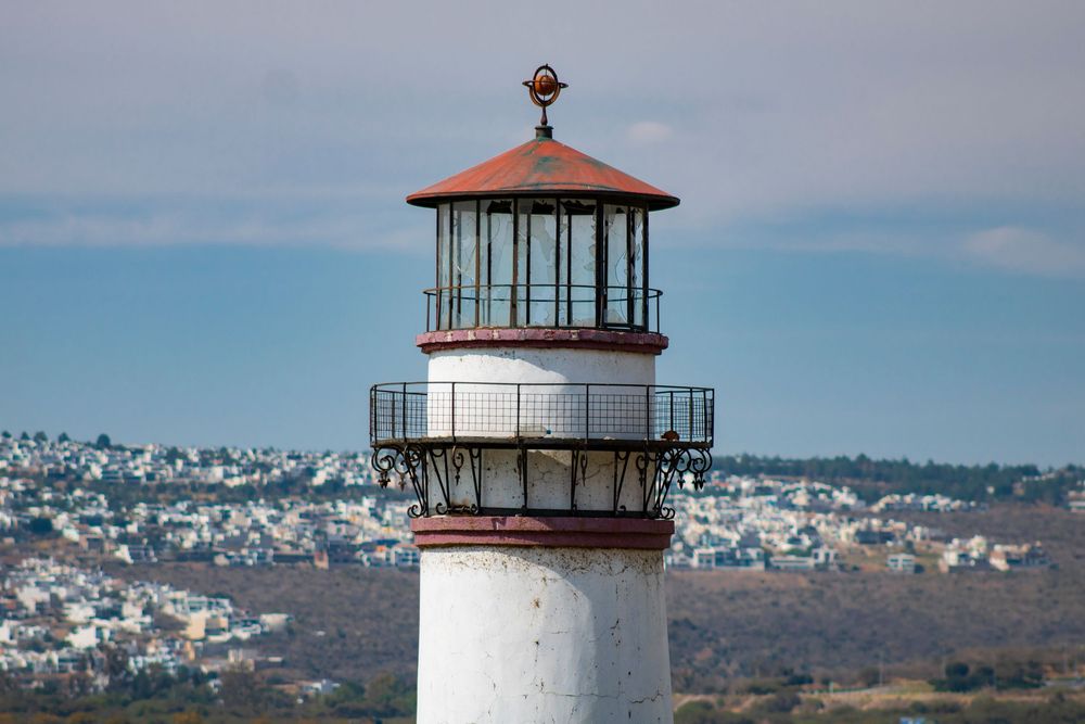 White and red lighthouse tower with balcony against blue sky with coastal city in the background.