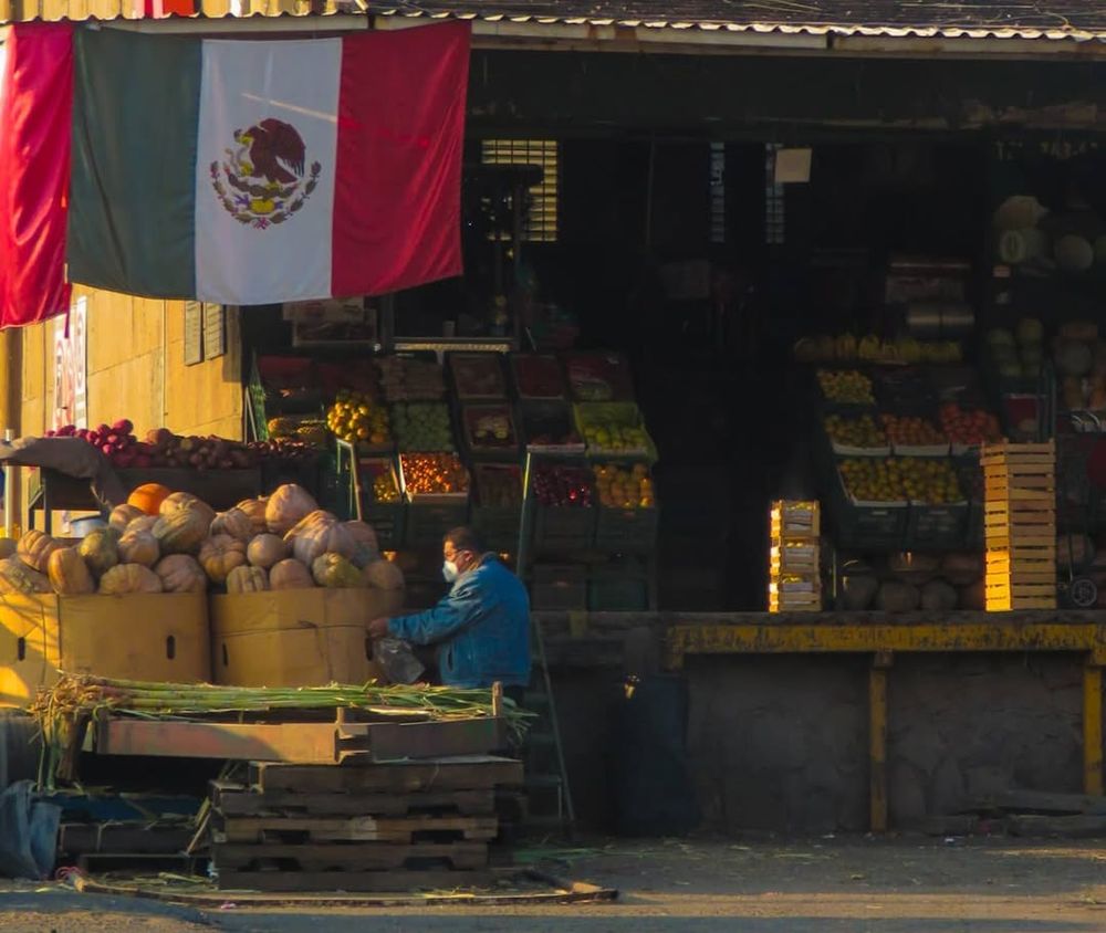 Mexican fruit market stall at golden hour with large Mexican flag hanging above fresh produce.