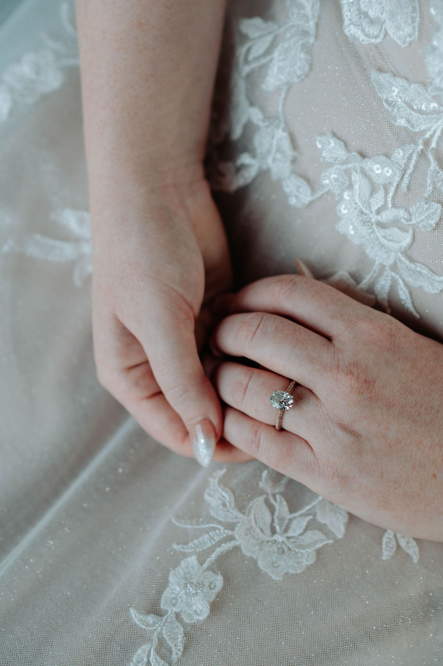 Close-up of hands showing an engagement ring against delicate lace wedding dress fabric.