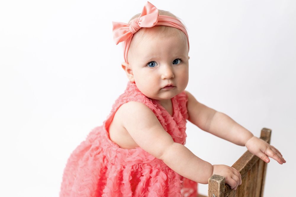 Cedar park TX photography milestone session, baby girl in white crib, coral pink dress and bow in her hair, studio White