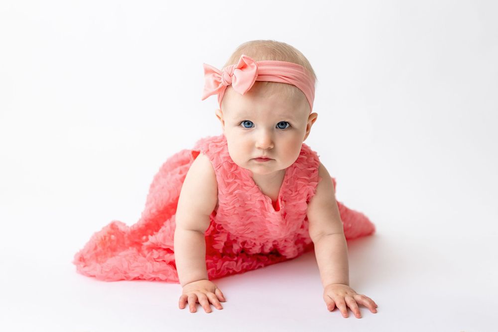 Cedar park TX photography milestone session, baby girl in white crib, coral pink dress and bow in her hair, studio White