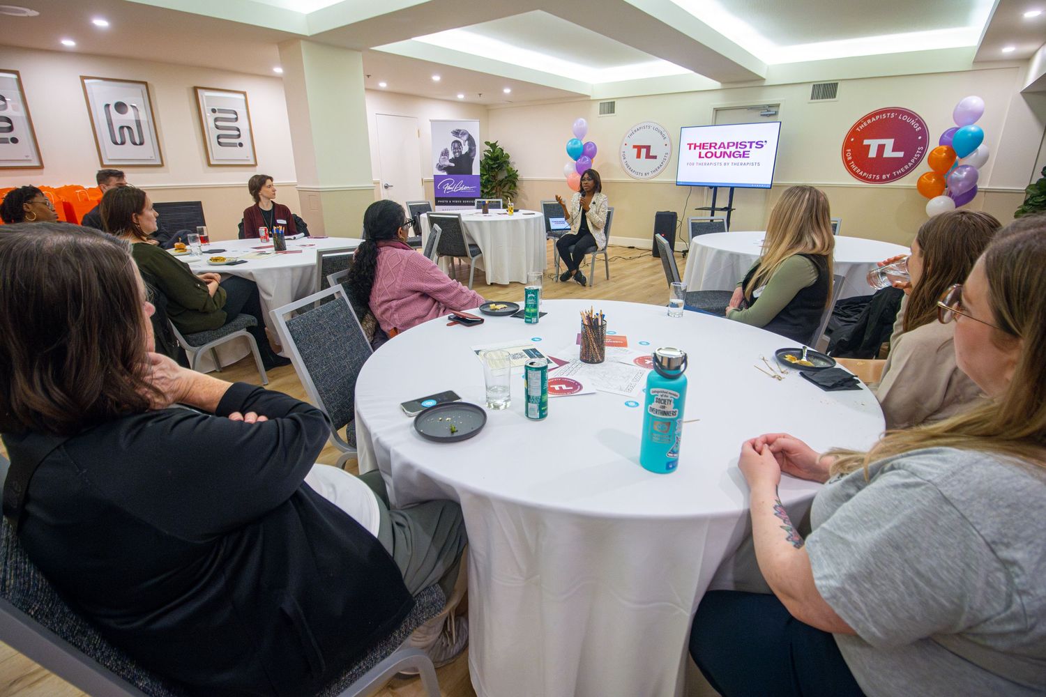 Group seated at round tables in a conference room with presentation screen and banners displayed in the background.