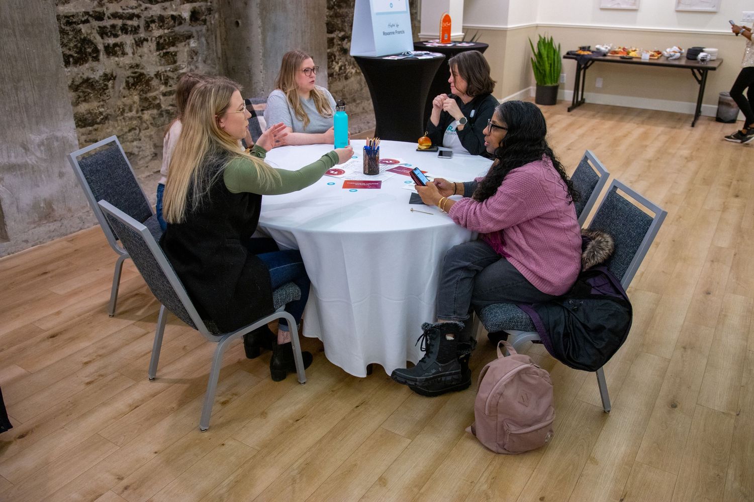 A group of people sitting at a round table having a discussion in a room with wooden floors and stone walls.
