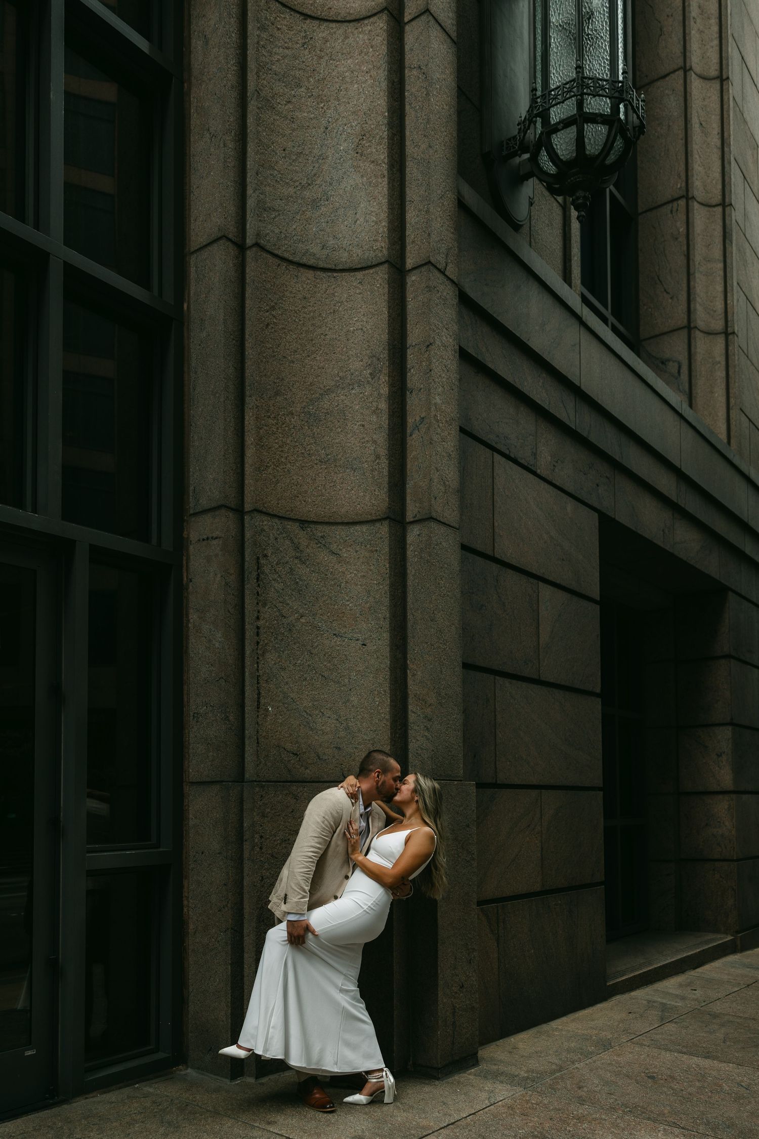 A couple in wedding attire embrace against a dramatic stone building wall.