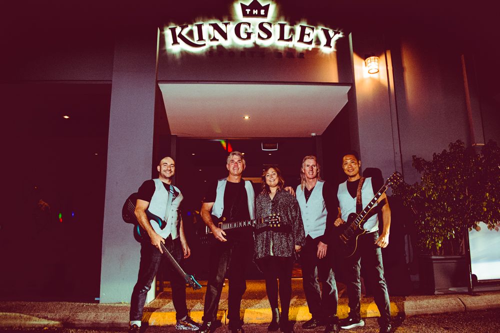 A music group poses together at night in front of the King's Head venue sign glowing above them.