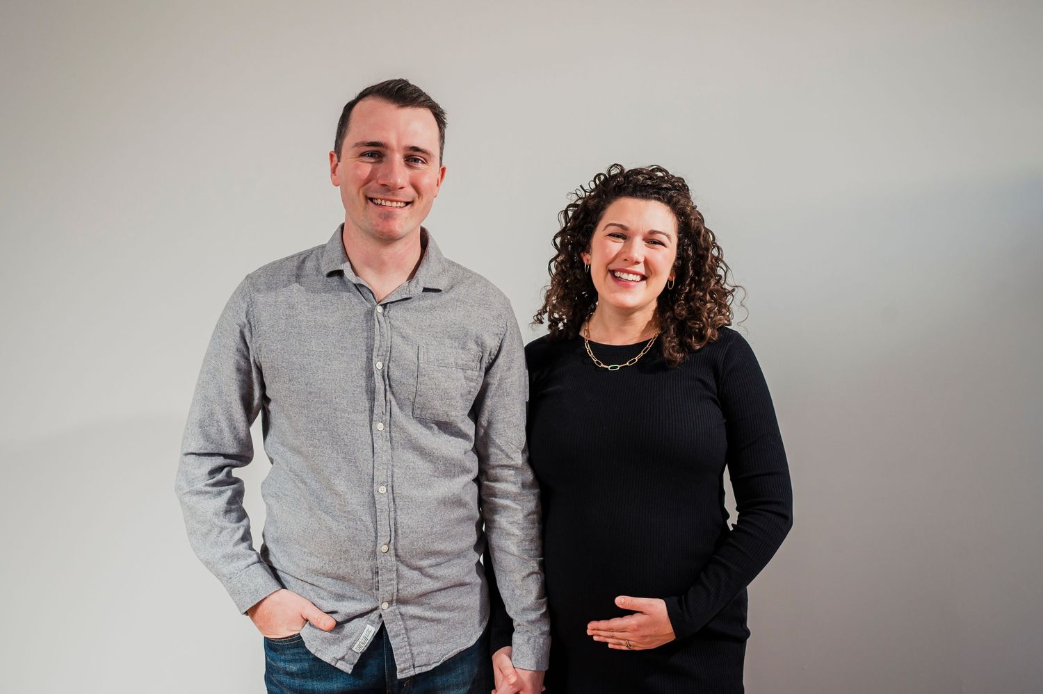 Couple standing together against a light gray background, one wearing a gray button-up and the other in a black sweater.