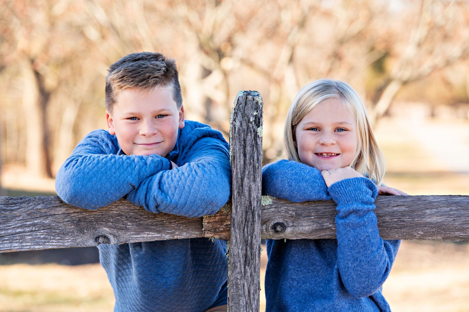 Two children in matching blue sweaters lean against a rustic wooden fence outdoors during autumn.