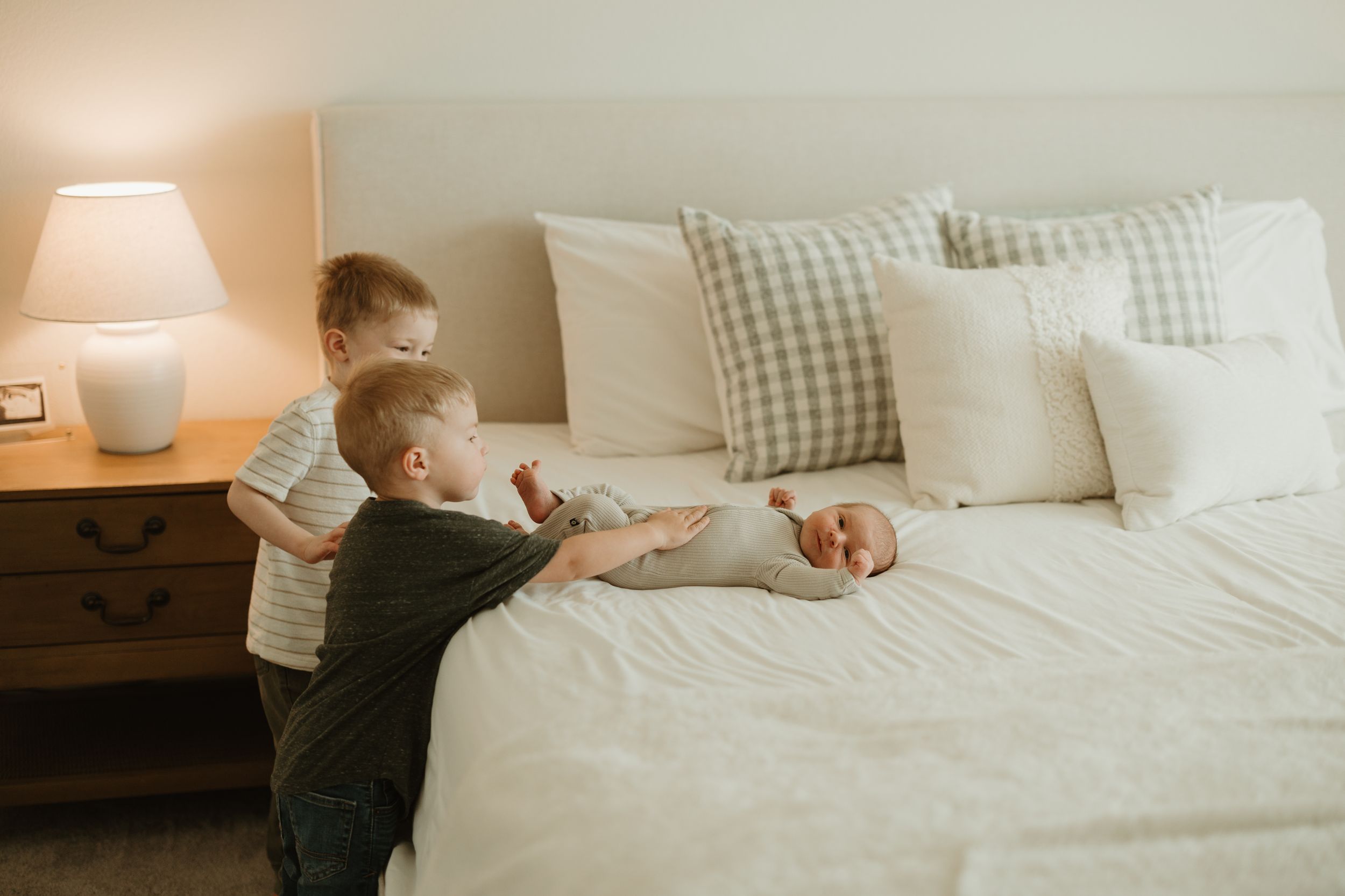 Kids playing on a cozy white bed with plush pillows and a gingham pattern headboard next to a bedside table with lamp.