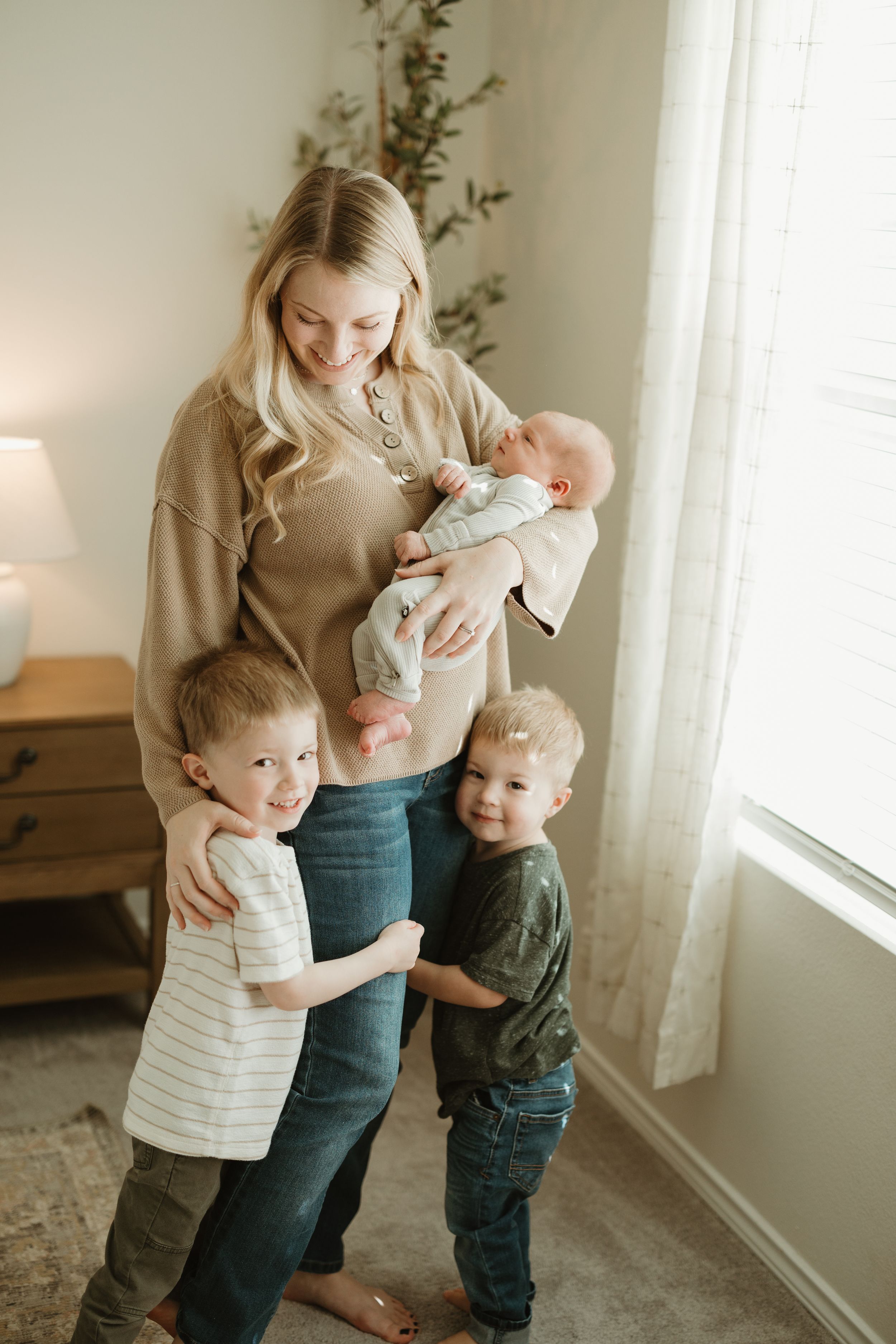 Gentle family moment by a bright window as young children embrace their parent holding a newborn in a cozy living room.