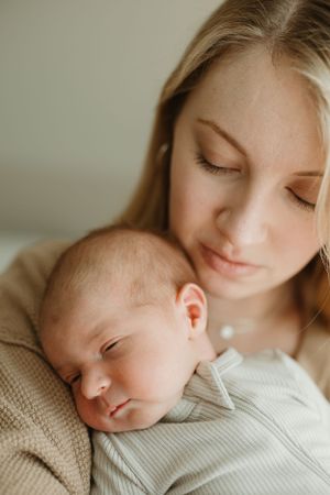 A newborn baby sleeps peacefully against a woman wearing a beige sweater in a tender moment.