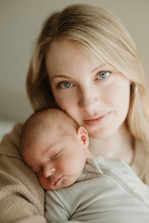 A tender moment captured as a sleeping newborn rests against someone wearing a beige sweater in soft natural light.