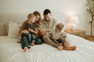 A family sits together on a white bed, looking at devices and sharing a cozy moment in a warmly lit bedroom.