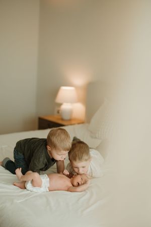 Two young children sharing a tender moment with their sibling on a white bed next to a table lamp.