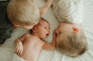Three young children lying together on white bedding in an intimate bonding moment captured from above.