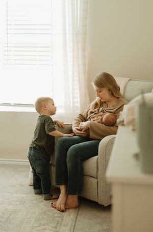 A cozy living room scene shows a toddler visiting during feeding time near a window with soft light streaming in.