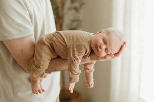 A sleeping baby in a brown onesie rests peacefully in someone's arms against a light-colored background.