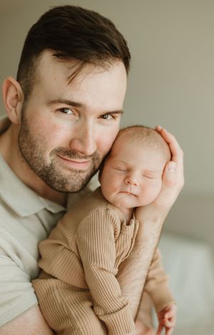 A tender portrait shows an adult holding a sleeping newborn baby wearing a brown knit outfit against a neutral background.