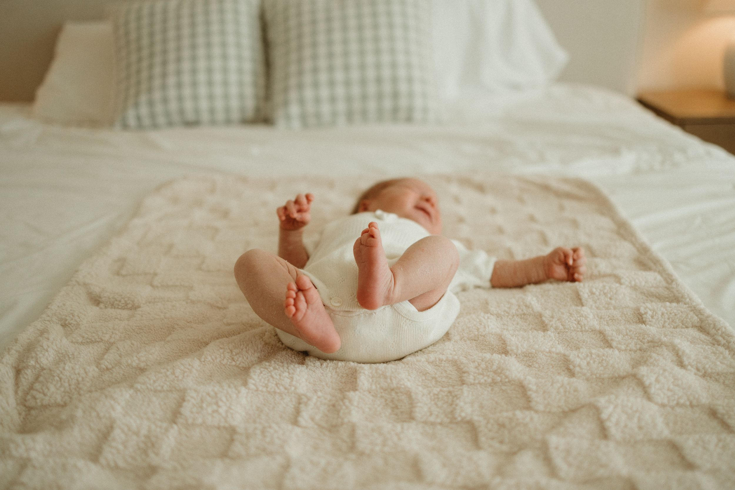 Baby wearing white clothing lies on their back on a textured beige blanket with gingham pillows in background.