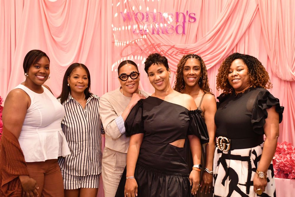 A group of people wearing black and white outfits pose together at a social event with a pink backdrop.