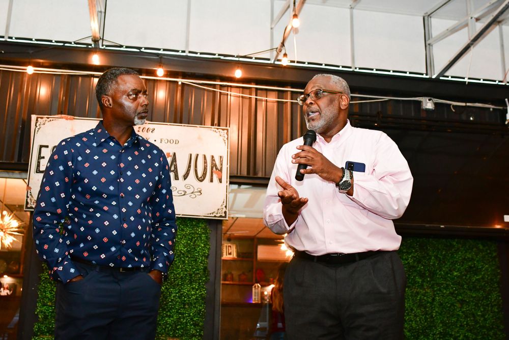 Two people speaking together on stage, one wearing a blue polka dot shirt and one in pink dress shirt.