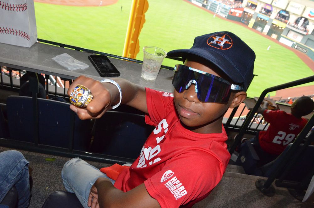 Young baseball fan poses wearing sunglasses and team gear in stadium seating.