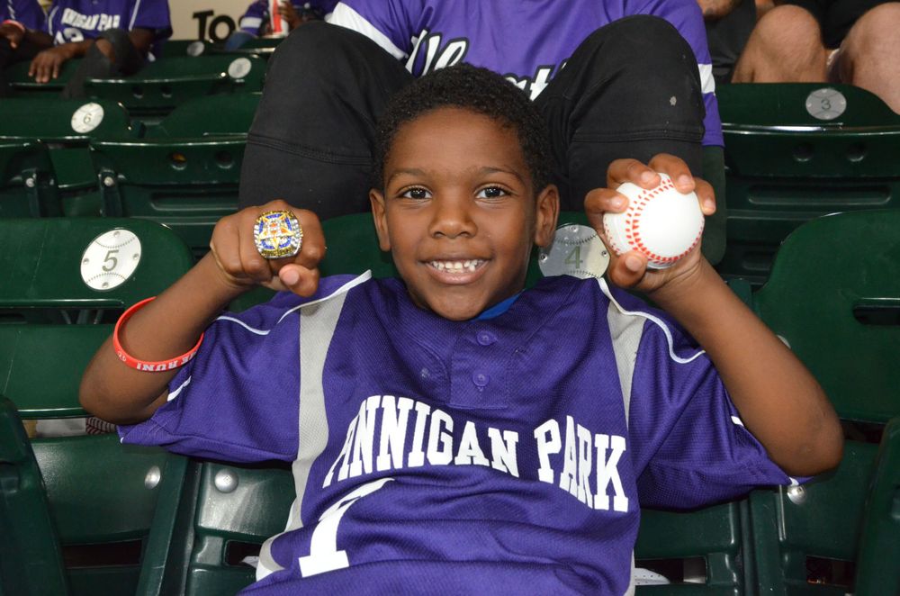 Young fan in purple shirt shows off baseball souvenirs at the ballpark.