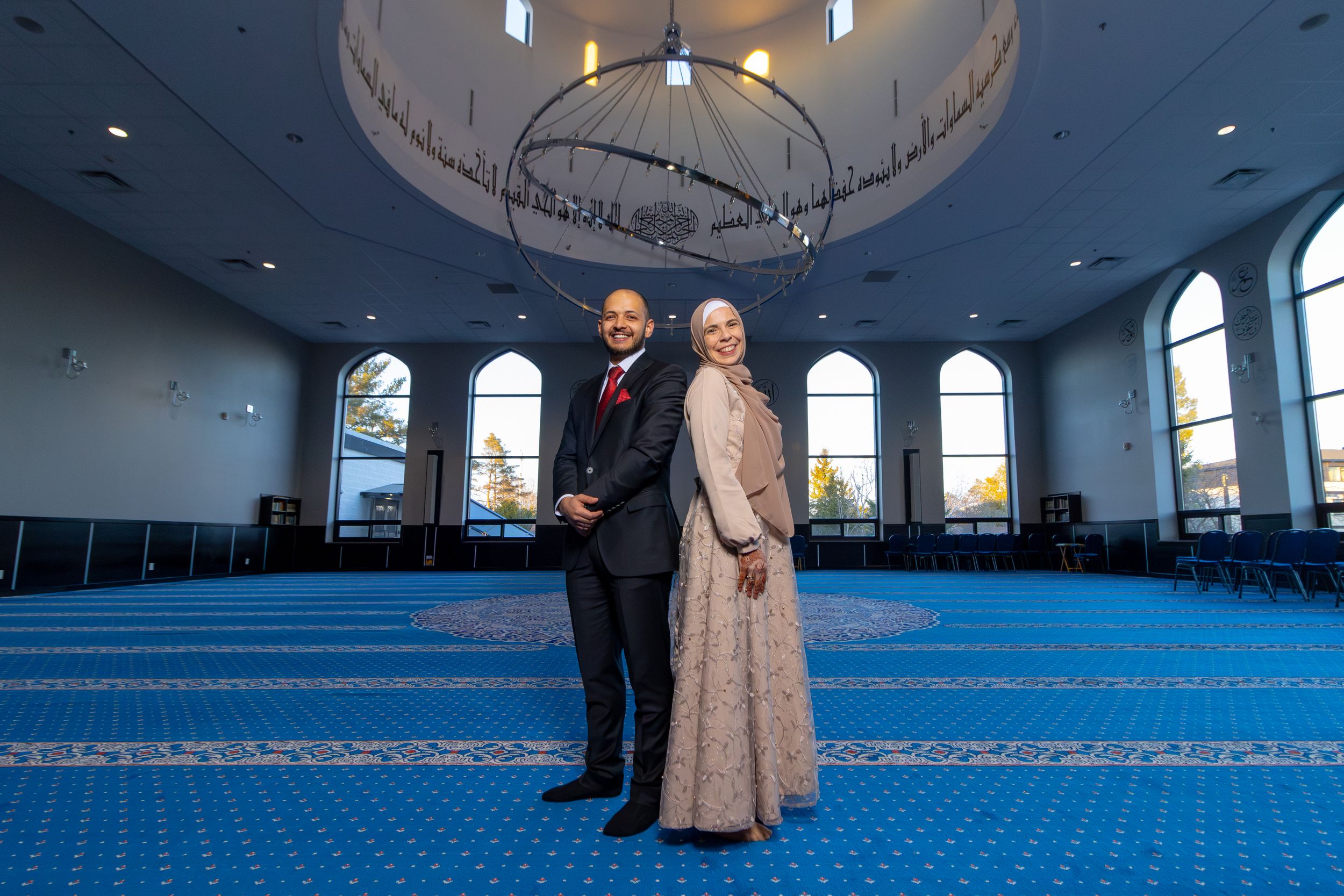 Two people stand together posing inside a spacious mosque with blue carpeting and arched windows.