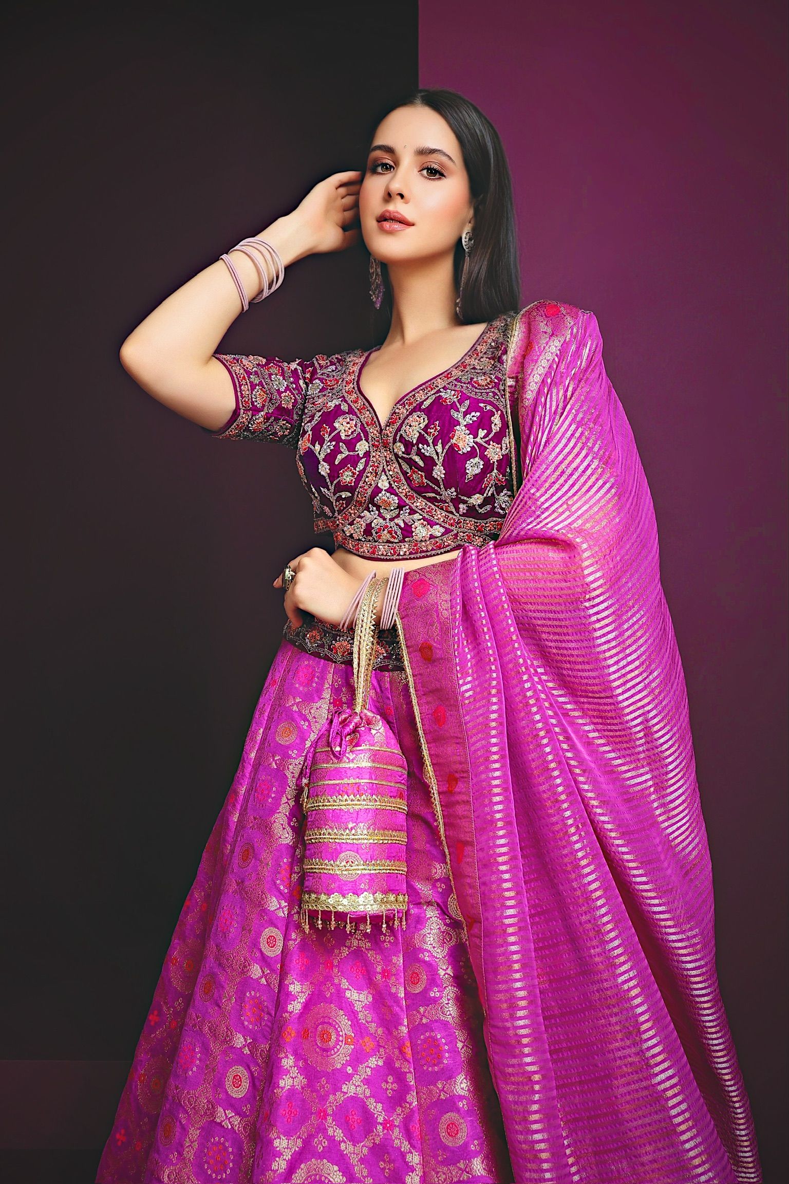 Woman in vibrant pink lehenga with purple embroidered blouse and dupatta, holding potli bag, posing elegantly.