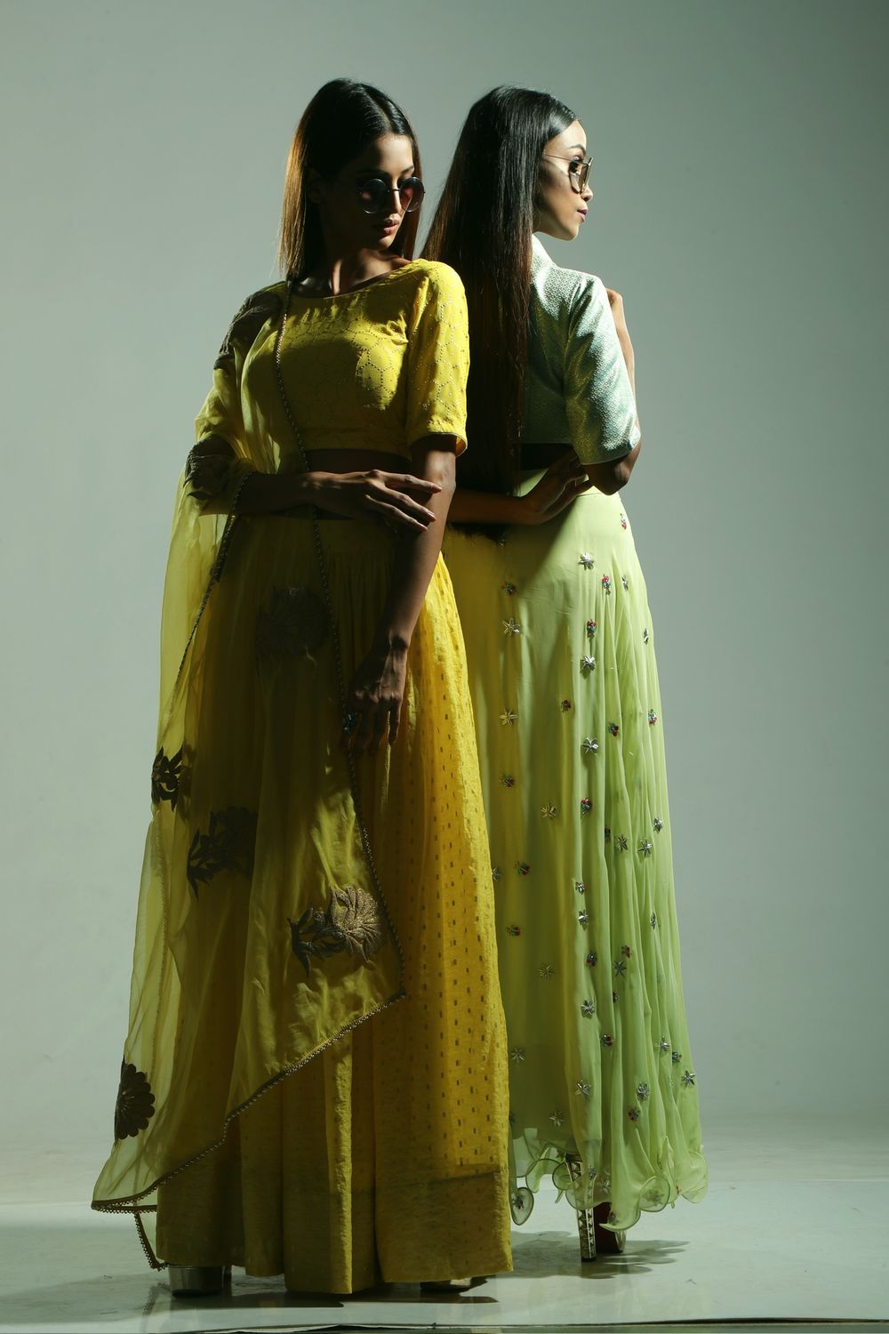Two women in elegant yellow and green Indian lehengas pose back-to-back in a fashion editorial shoot.