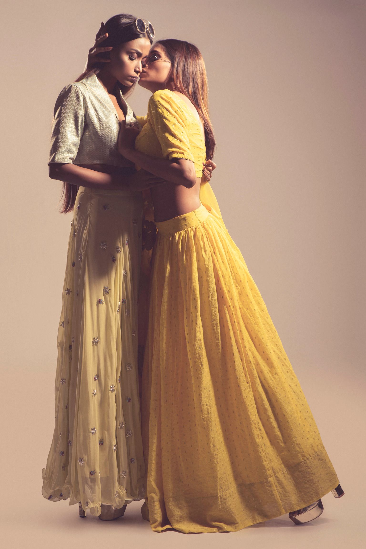 Two women in elegant Indian lehengas, one in white and one in yellow, posing together in a studio setting.