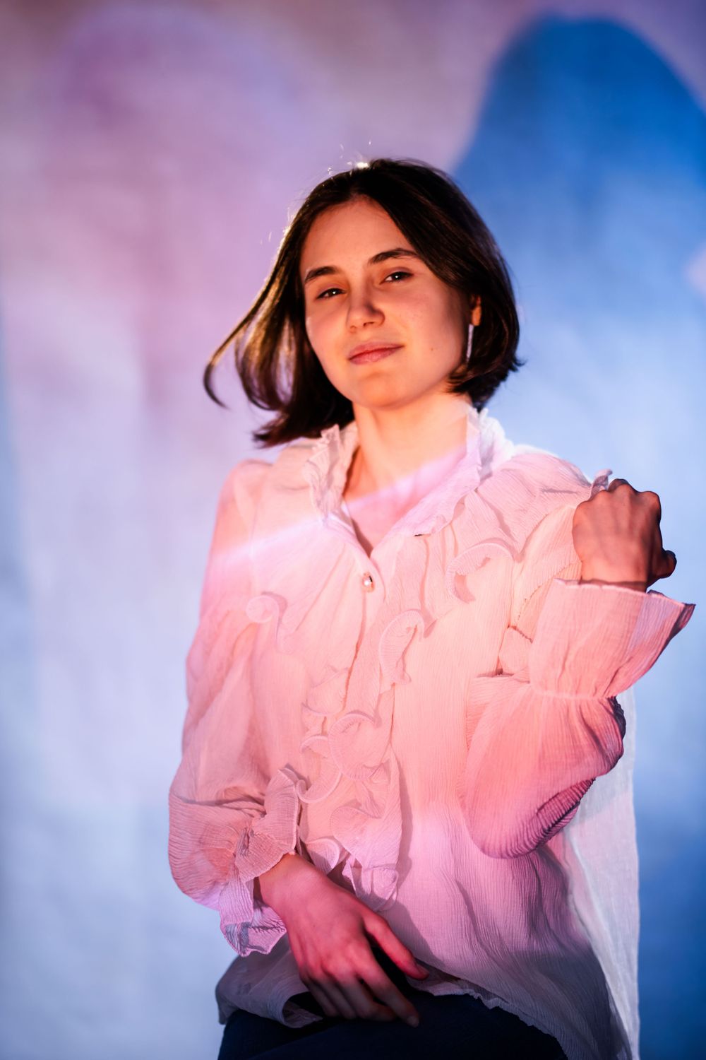 Portrait against blue and purple background wearing light pink button-up blouse in vibrant lighting.