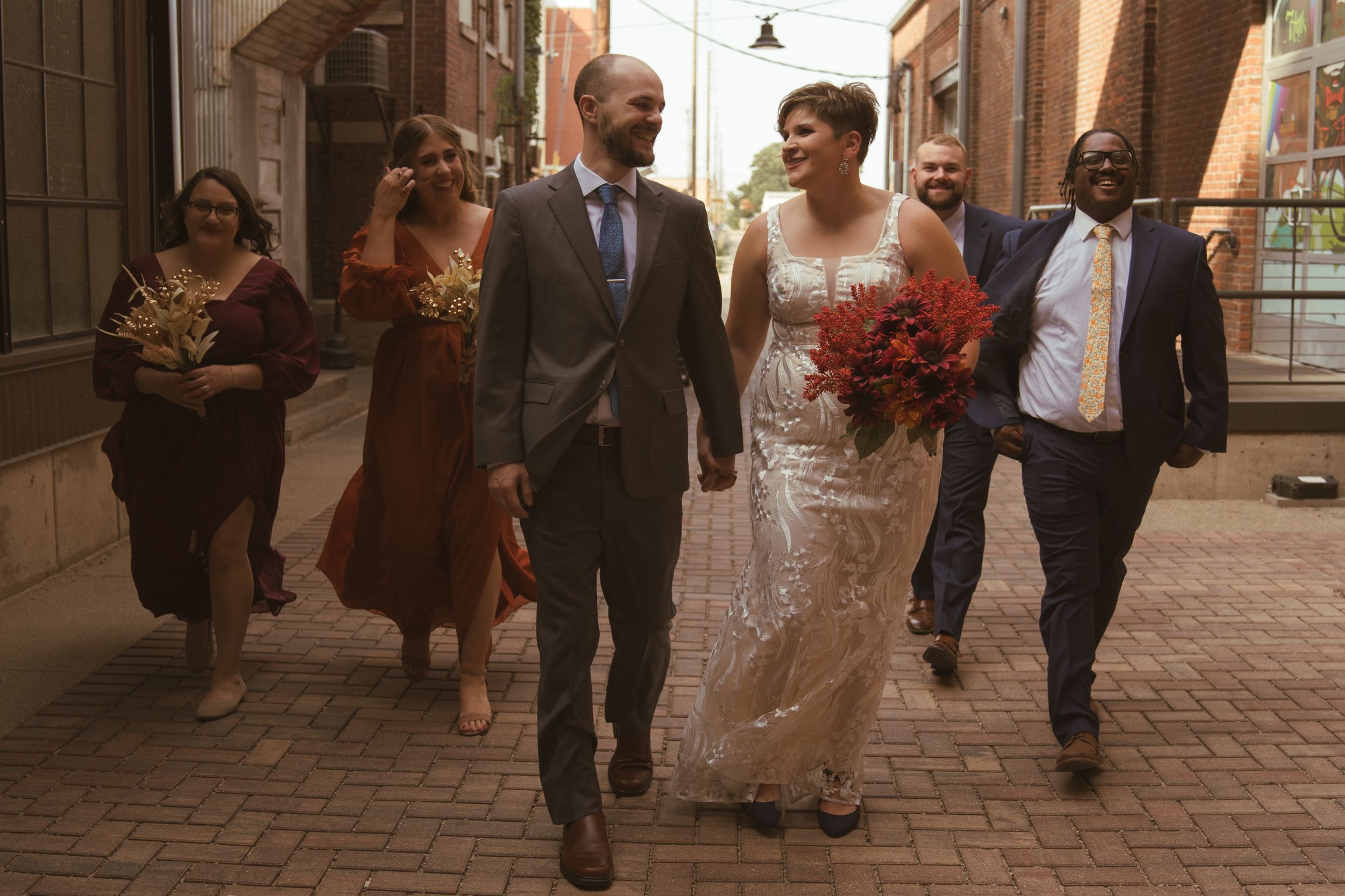 Wedding party walking and laughing together down an urban alley with brick buildings.