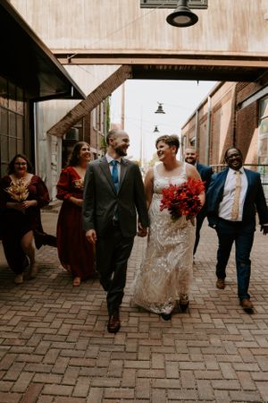 A wedding procession walks through an urban setting with red floral accents.