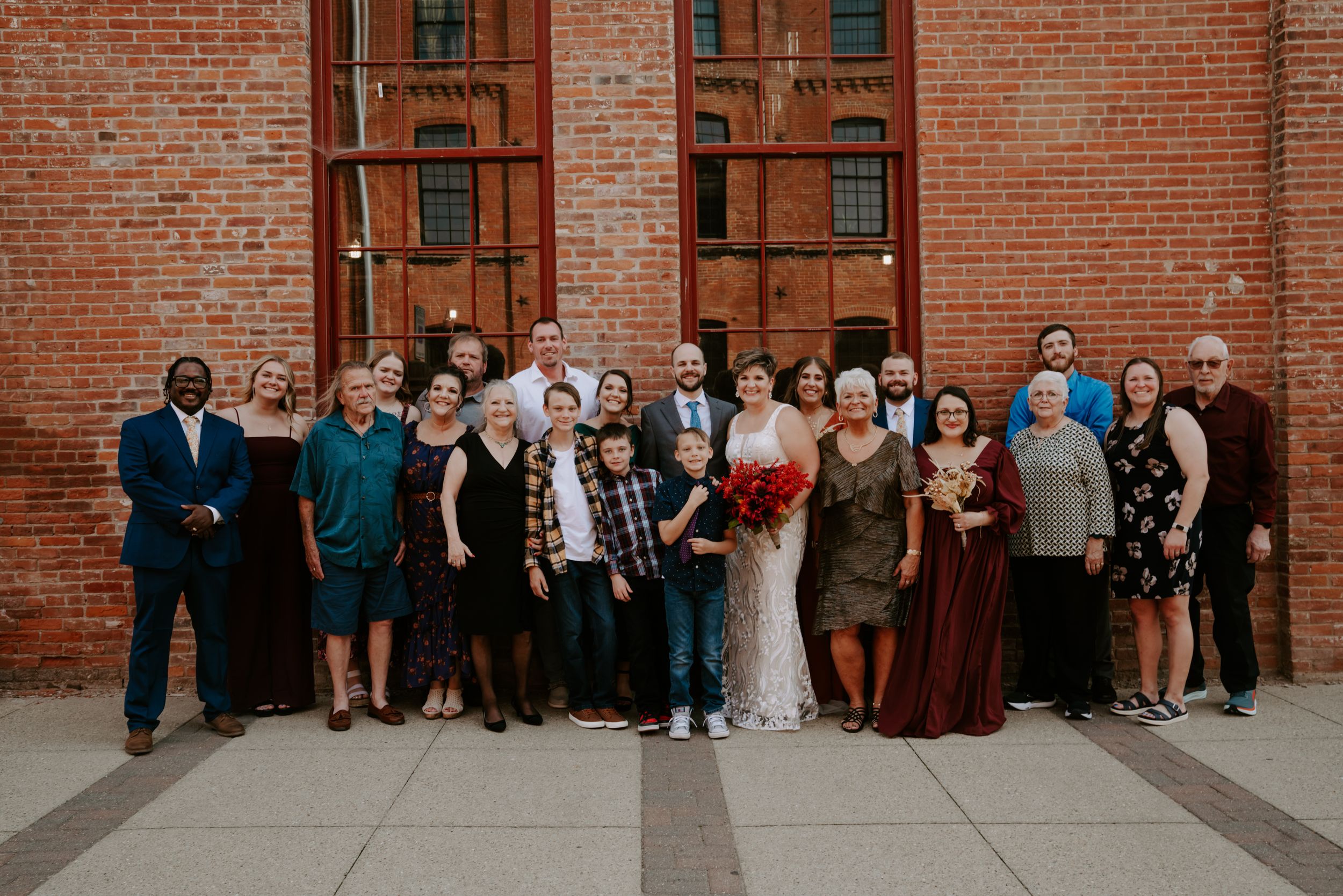 Large wedding group photo against rustic brick wall backdrop.
