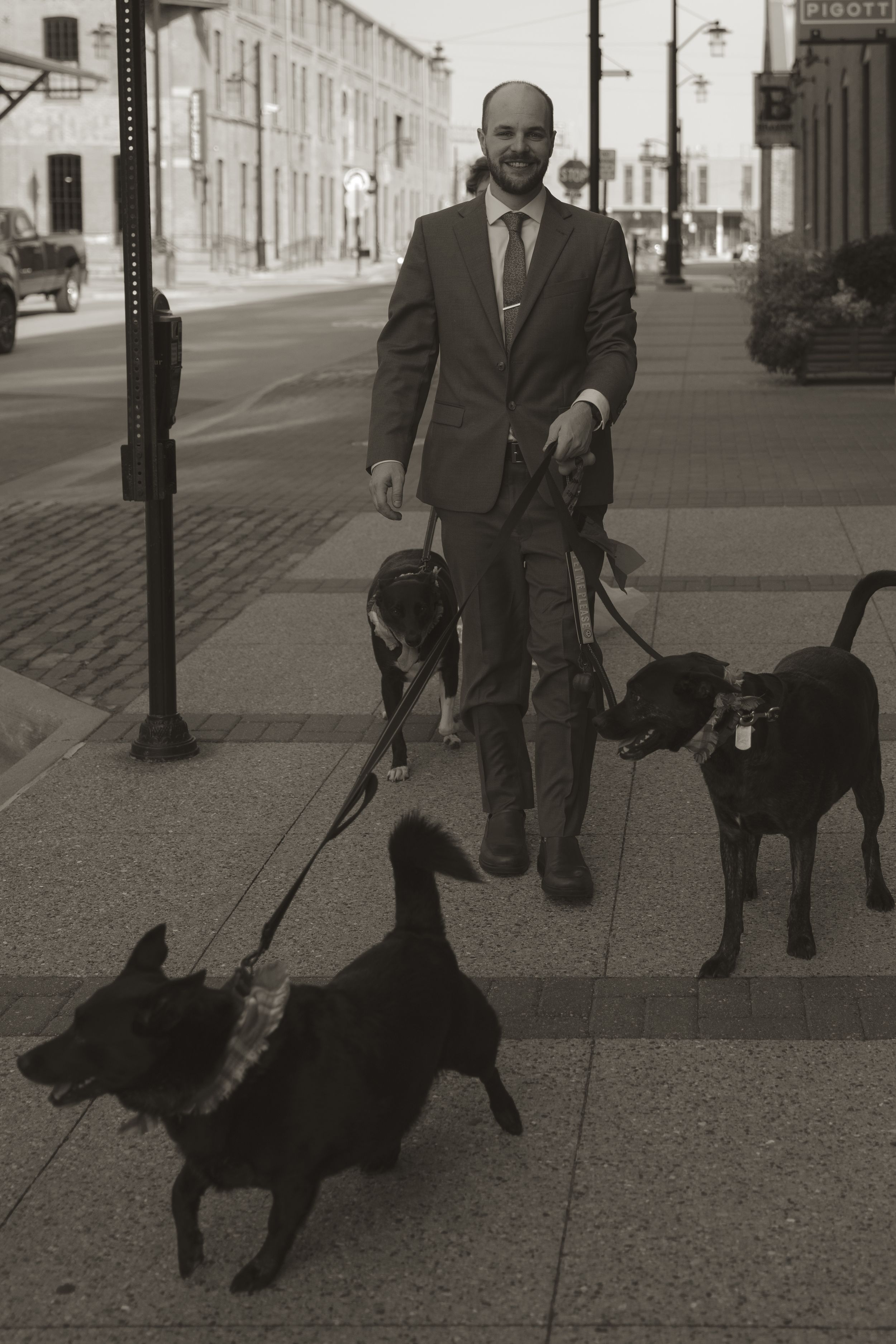 A person in formal attire walks a black dog on a city sidewalk in black and white.