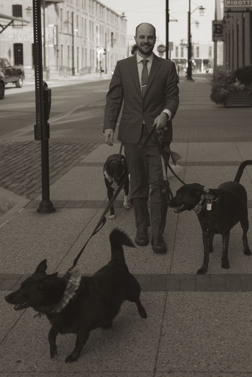 A person in formal attire walks a black dog on a city sidewalk in black and white.