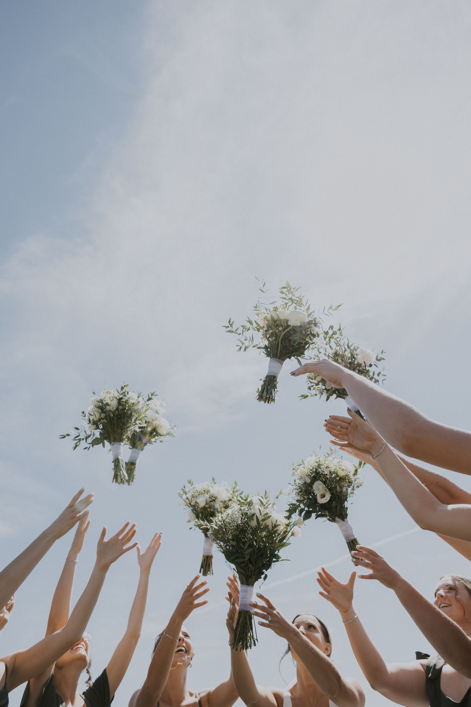 Multiple hands reach upward toward white floral bouquets being tossed against a bright blue sky.