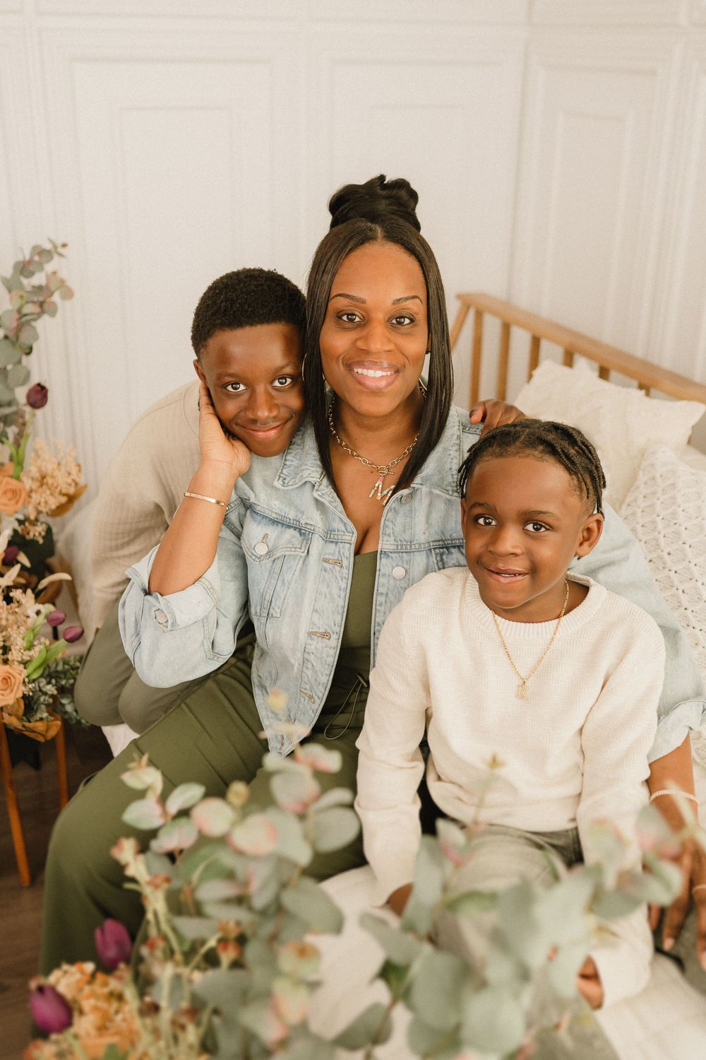 A family portrait showing three individuals posing together surrounded by floral arrangements and soft decor.