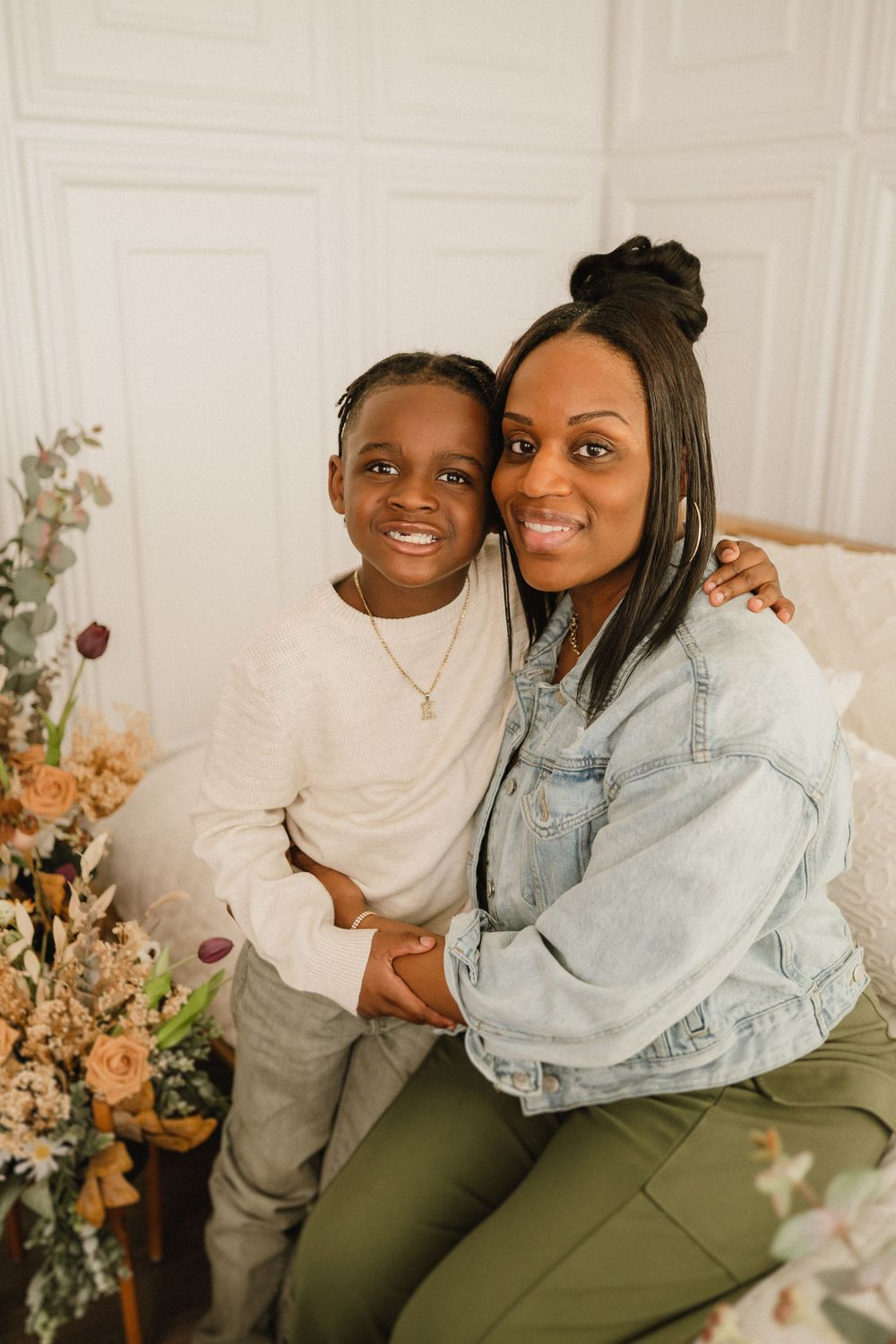 Mother and child pose together in front of floral arrangement wearing casual denim and white clothing.