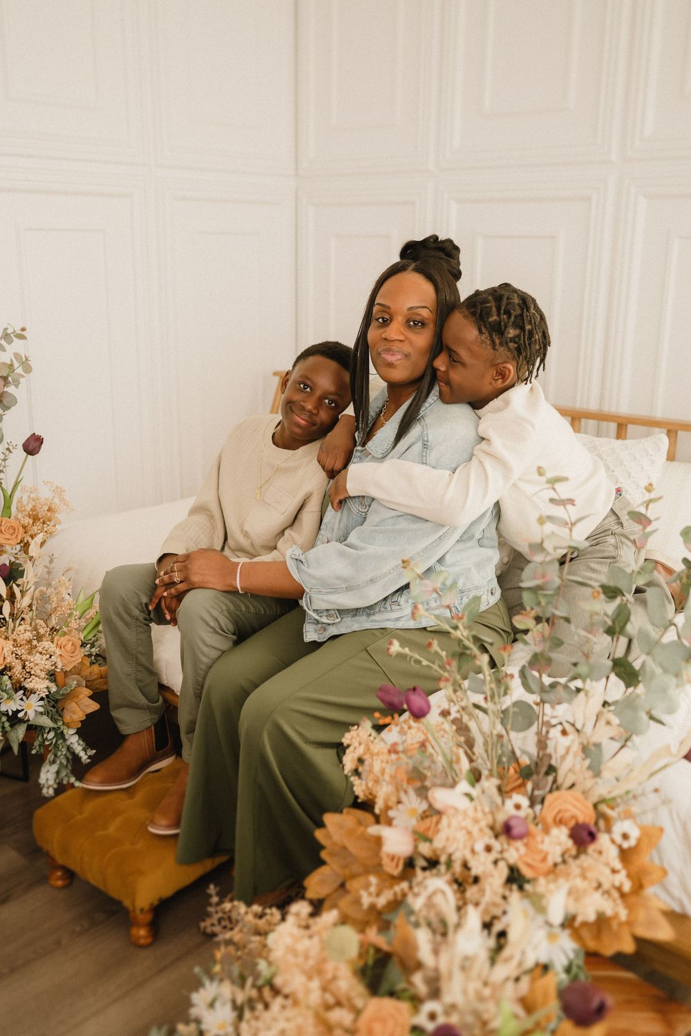 A loving family sits together on a wooden ottoman surrounded by peach and pink floral arrangements in a bright room.