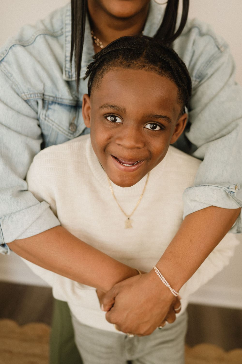 A young child wearing a white sweater and necklace smiles brightly at the camera in a casual indoor portrait setting.