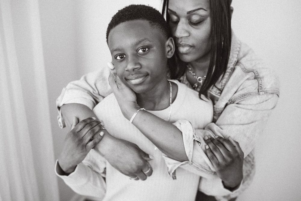 Black and white portrait of parent lovingly embracing child from behind in tender moment against light background.