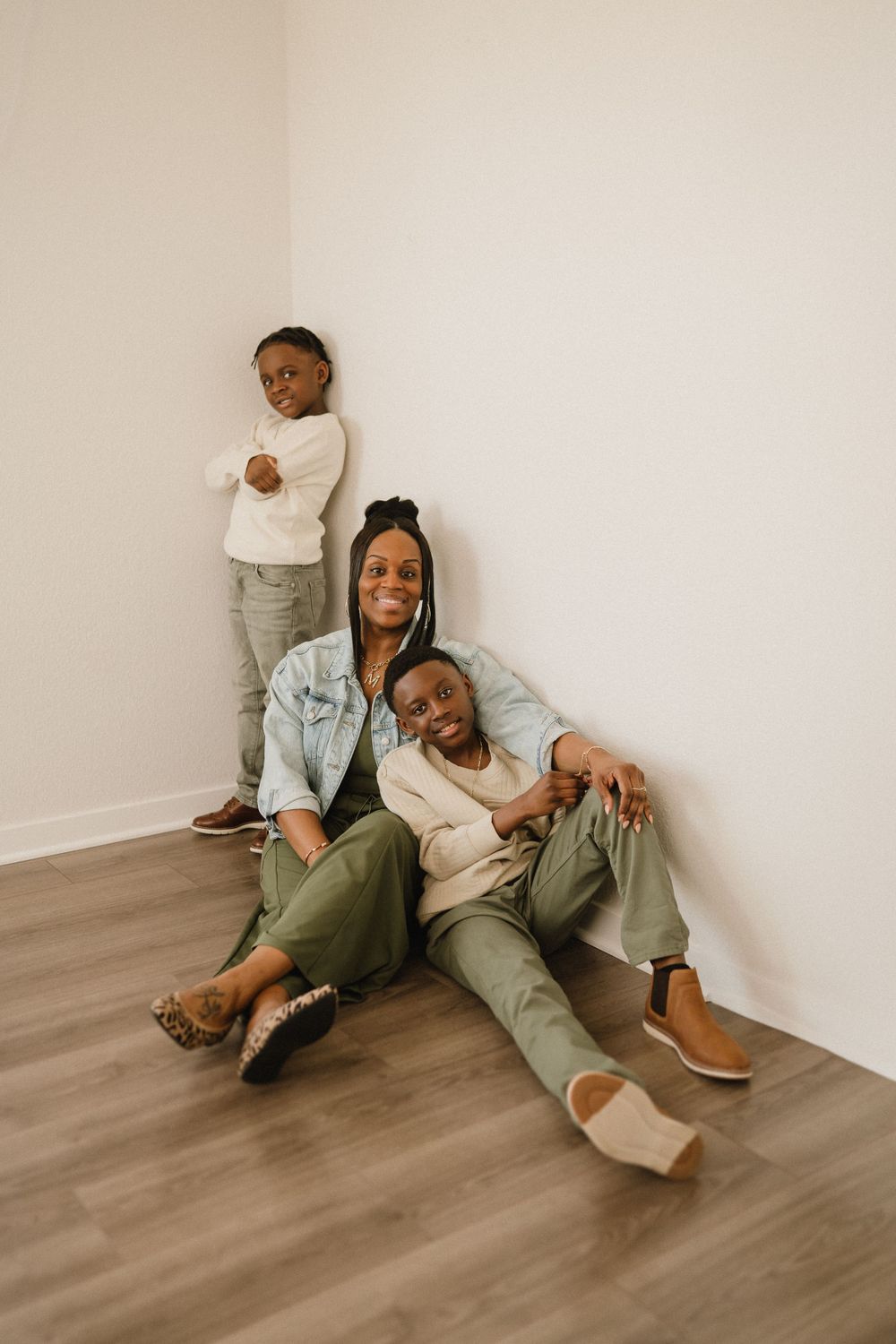 A family sits together on a hardwood floor against a beige wall in casual stylish outfits.