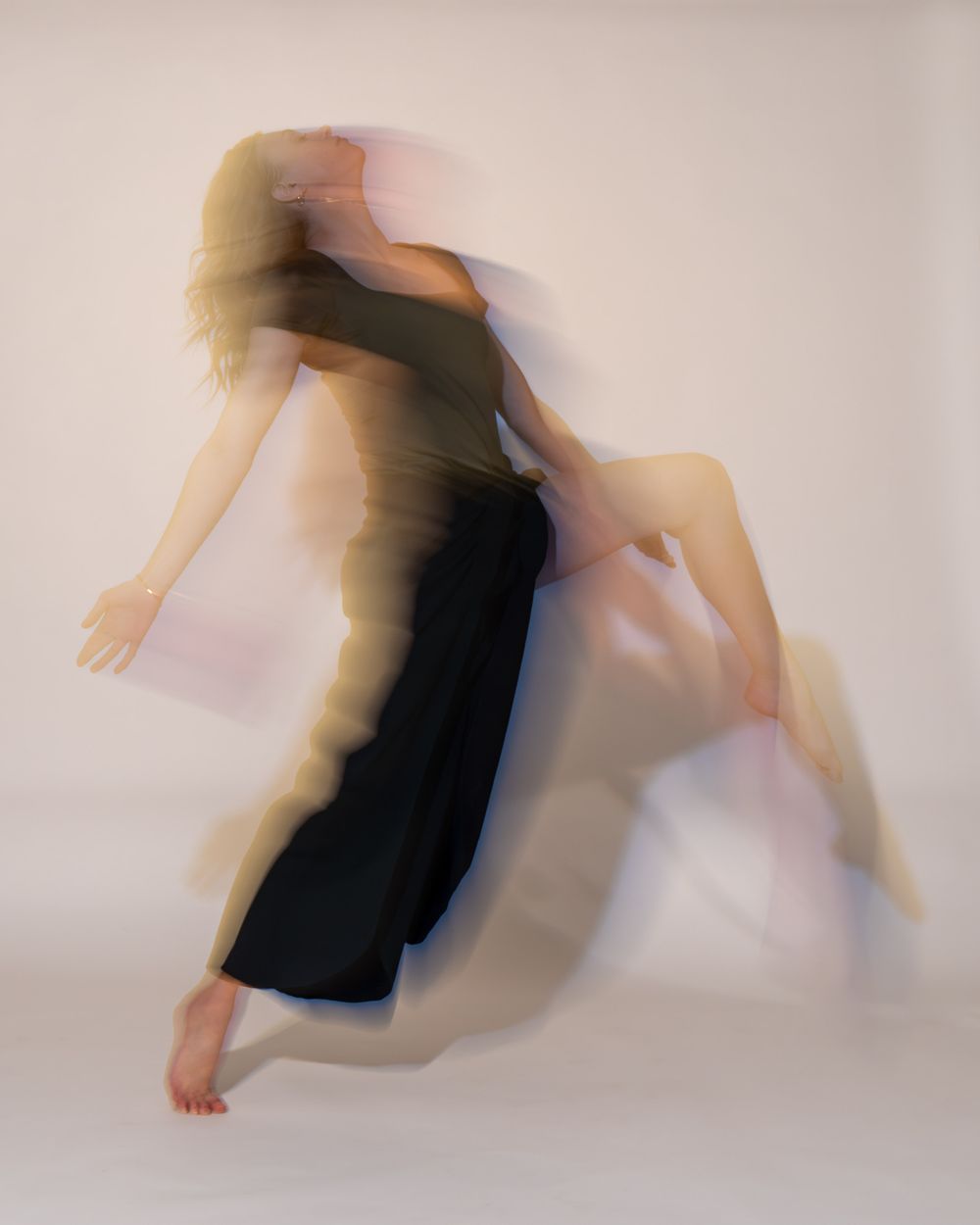 Artistic motion blur photograph of a figure in a black dress leaning back in a graceful dance-like pose against white backdrop.